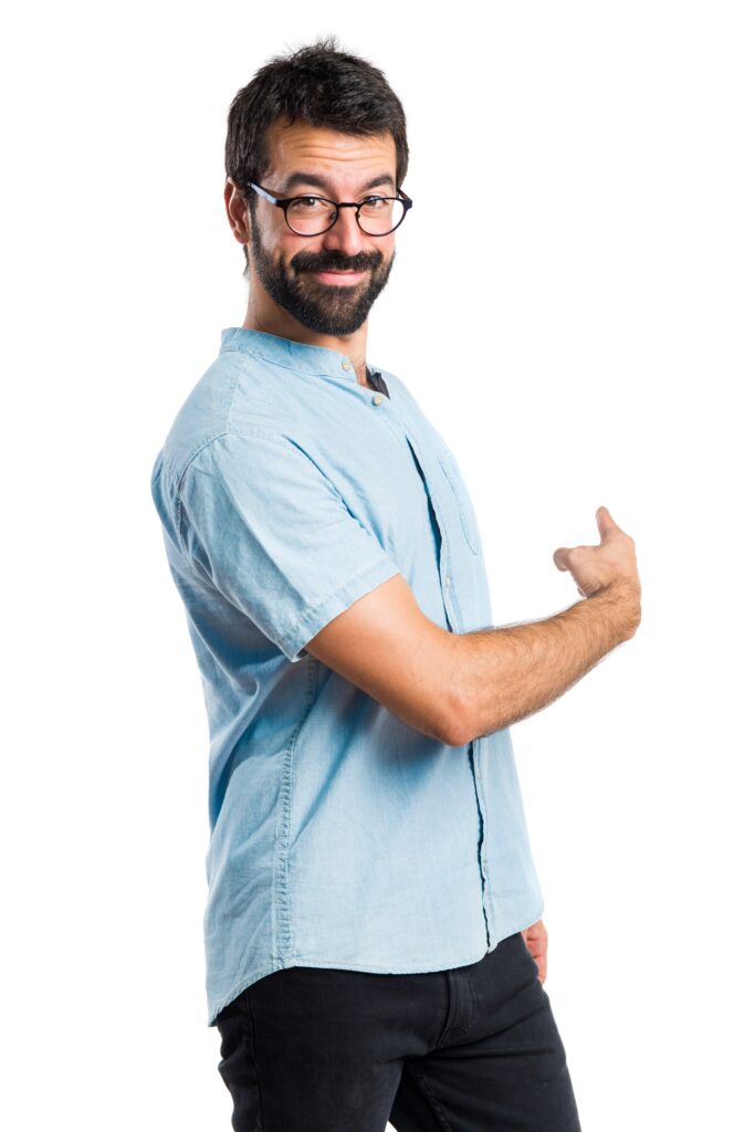 Man with dark hair, beard and round glasses wearing a light blue shirt, smiling slightly, and pointing backward against a white studio background.